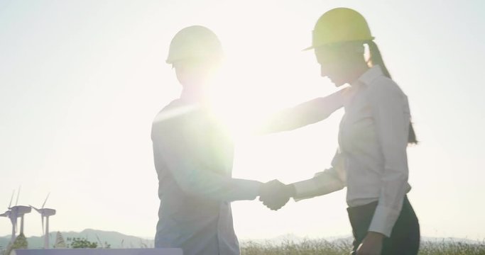 Two engineers talk about the project for the construction of a renewable energy wind farm; the two shake hands with the finished work in a wind farm at sunset.