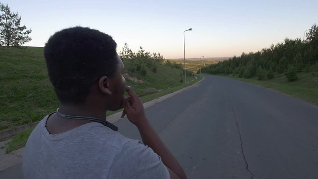 Rear view of young african-american man walking down the empty road and smoking cigarette at sunset