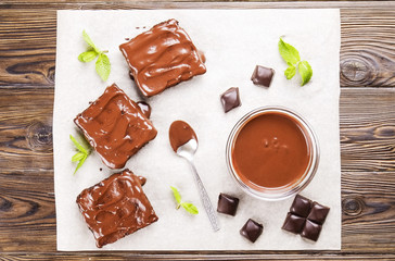 Square pieces of homemade brownies, dark chocolate spread paste, spoon and mint on white baking paper. High calorie pastry on table. Cocoa based sweets, bad for figure. Close up, background, top view.