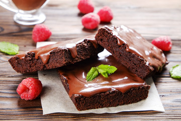 Square pieces of homemade brownies, dark chocolate spread paste, raspberry & mint leaf on wood table. High calorie pastry. Cocoa based sweets, bad for figure. Close up, copy space background, top view