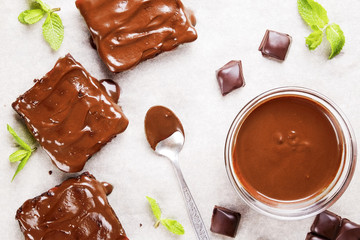 Square pieces of homemade brownies, dark chocolate spread paste, spoon and mint on white baking paper. High calorie pastry on table. Cocoa based sweets, bad for figure. Close up, background, top view.