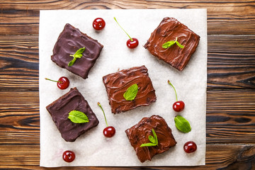 Square slices of homemade brownies, dark chocolate spread paste, cherry & mint leaves on wood table. High calorie pastry. Cocoa based sweets, bad for figure. Close up, copy space background, top view.