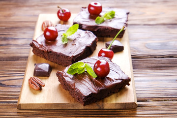 Square pieces of homemade brownies, dark chocolate spread paste, cherry & pecan nut on wooden table. High calorie pastry. Cocoa based sweets, bad for figure. Close up, copy space background, top view.