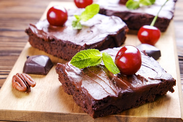 Square pieces of homemade brownies, dark chocolate spread paste, cherry & pecan nut on wooden table. High calorie pastry. Cocoa based sweets, bad for figure. Close up, copy space background, top view.