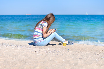 Fototapeta na wymiar Young beautiful woman enjoys relaxing at the sea. She sits on the sand on a blue sea background with a smartphone in her hands