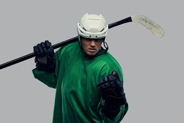 Angry professional hockey player in green sportswear standing with a hockey stick on a gray...