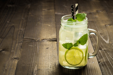 Cold Mojito Cocktail with Ice, Lemon and Mint Leaves in Mason Jar on Rustic Dark Wooden Background as Summer Concept