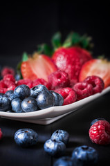 Blueberries, raspberries and strawberries, strawberries in a cut on a white plate on a black background