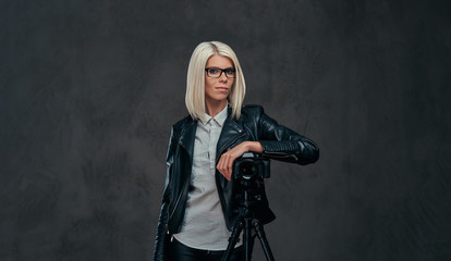 Smart blonde female photographer in trendy clothes posing while leaning on a professional camera with a tripod at a studio, looks at the camera.