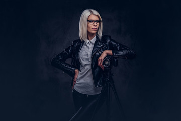 Fototapeta na wymiar Smart blonde female photographer in trendy clothes posing while leaning on a professional camera with a tripod at a studio, looks at the camera.