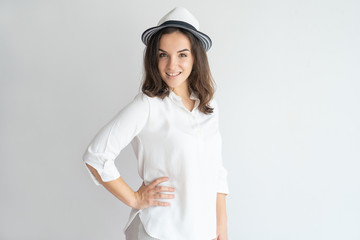 Stylish positive girl in summer hat posing at camera. Trendy young Caucasian woman in white shirt and round hat keeping hand on hip and smiling. Fashion and summer outfit concept