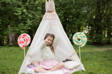 Little girl lying and playing in a tent, children's house wigwam in park. Happy mothers day. Child with big donuts and heart-shaped pillows. Summer, childhood concept. © MartaKlos