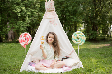 Little girl lying and playing in a tent, children's house wigwam in park. Happy mothers day. Child with big donuts and heart-shaped pillows. Summer, childhood concept. © MartaKlos