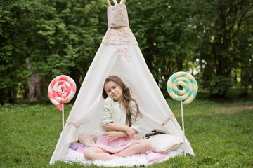Little girl lying and playing in a tent, children's house wigwam in park. Happy mothers day. Child with big donuts and heart-shaped pillows. Summer, childhood concept. © MartaKlos