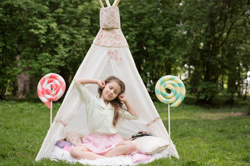 Little girl lying and playing in a tent, children's house wigwam in park. Happy mothers day. Child with big donuts and heart-shaped pillows. Summer, childhood concept. © MartaKlos