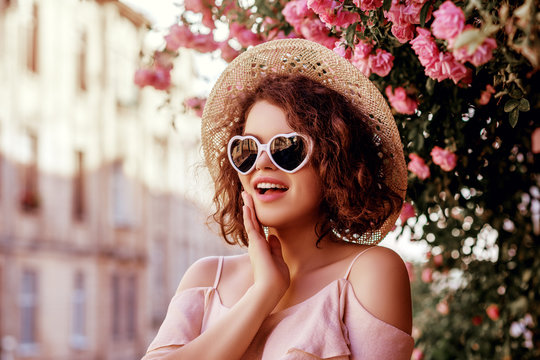 Outdoor close up portrait of young beautiful surprised curly girl wearing stylish heart sunglasses, straw hat, pink blouse. Model posing near blooming roses. Summer fashion concept. Copy space
