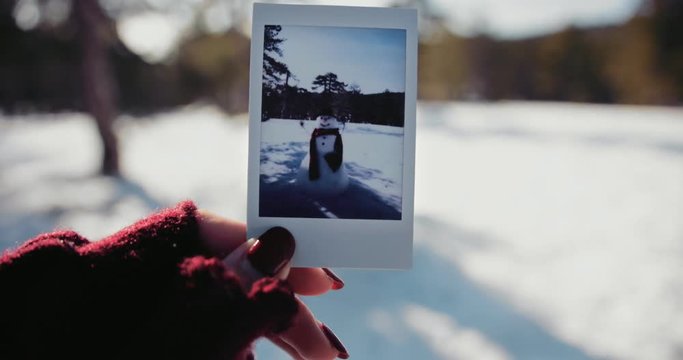 Woman holding polaroid photo of snowman in snowy mountain forest