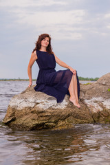 Young attractive girl in a long black dress sitting on a rock by the river.