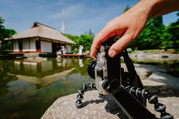 Photograph male hand handling an old vintage camera on tripod in front of water pond with landmark japanese house and wedding couple in Planten un Blomen Park Hamburg, Germany