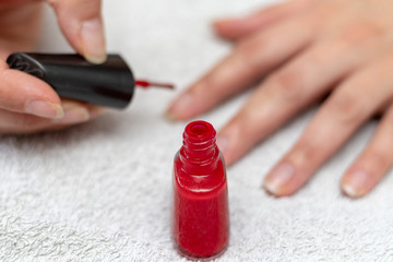Small bottle of red nail enamel in front of the hands of a woman coating the nails with red lacquer