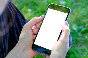 The girl is sitting on the grass and looking into the smartphone holding it in her hands. Mockup image.