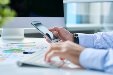 Crop shot of man in shirt using smartphone and computer working at desktop in light office. 