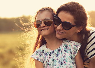 Happy fashion kid girl embracing her mother in trendy sunglasses and looking on nature background. Closeup toned portrait of happiness.