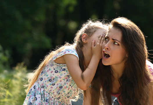 Happy Kid Girl Whispering The Secret To Her Surprising Shocked Mother With Open Mouth In Ear With Fun Face On Summer Green Tree And Grass Background.