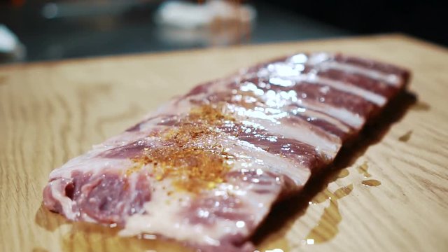 Young cook sprinkles meat fillets with seasonings in kitchen of bar. Professional strew pieces of pork with aromatic condiment on table in room. On wooden board is fresh meat, person sprinkles it with