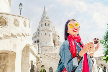 Fototapeta na wymiar Young asian woman tourist drinking frappe sweet cold coffee and tasty muffin at the Budapest city street near Fisherman Bastion. Travel in Hungary and fast food break concept