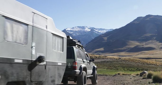 Van with trailer driving on gravel road, passes through badoo, stream. Surrounding of Varvarco valley, mountains with snow at background. Camera stays still while motorhome moves. Patagonia