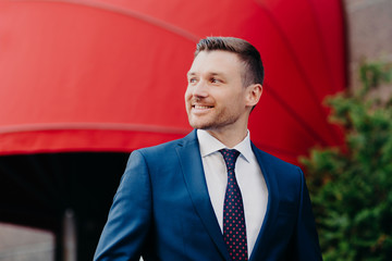 Outside view of handsome young male entrepreneur in formal suit, looks aside with glad expression, notices something pleasant, likes his work, stands outdoor. People, business, lifestyle concept