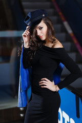 Fashion portrait of a pretty woman with naked shoulder, wearing a black peaked cap and elegant black dress. In hand blue leather jacket. Dark Background.
