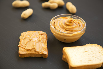 Bowl of peanut butter, toasts, unshelled peanuts over black background, side view. Close-up.