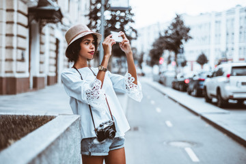 Young adorable black girl in the hat is taking photos of the street using her smartphone, with multiple cars next to her in defocused background and a copy space area on the right for text message