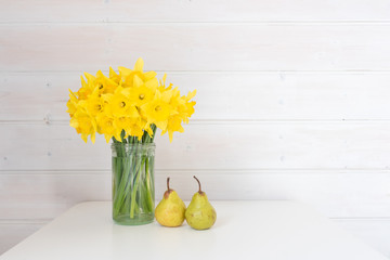 Yellow daffodils in glass jar on white table with two green pears against rustic white wood panelled wall
