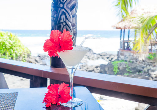 Alcoholic cocktail drink with red hibiscus flower garnish with wooden carving in background at beach bar on Upolu Island, Samoa, South Pacific