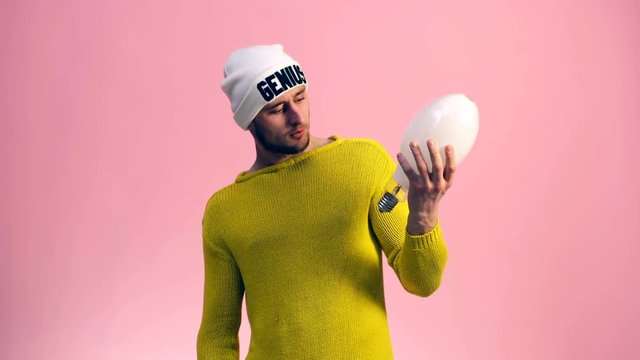 A man in a yellow sweater looks at a large white lamp on a pink background. Genius. Idea. Invention. Discovery