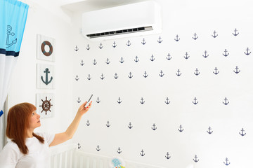Obraz premium Portrait Of A Happy Woman Holding Remote Control In Front Of Air Conditioner At Home in childrens room.Air conditioner inside the cute baby boy’s room.With hot weather,is a good solution for family.