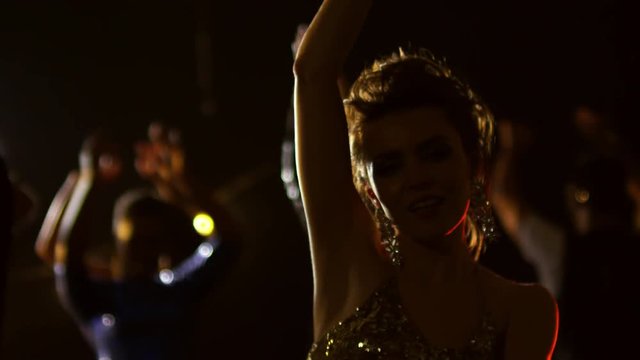 Medium shot of joyous young woman in sequin tank top dancing with hands in the air at music concert