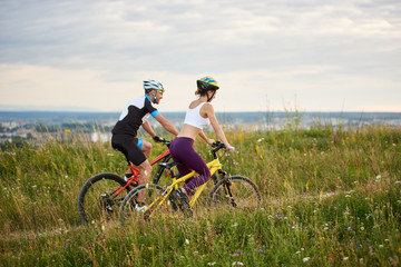 Obraz premium Two happy cyclists - man and woman riding on a hill in the grass with wild flowers, city and mountains in the distance. Both are dressed in sports clothes and helmets