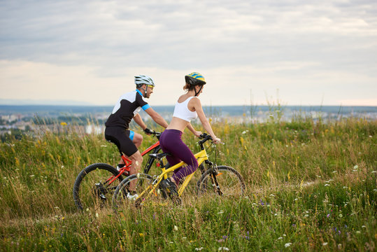 Two happy cyclists - man and woman riding on a hill in the grass with wild flowers, city and mountains in the distance. Both are dressed in sports clothes and helmets