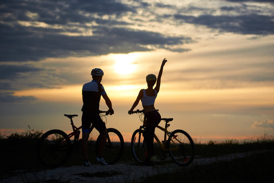 Rear view of a couple of cyclists standing with bicycles and enjoying the sunset. Man and woman are dressed in helmets and sportswear. Perfect sky with clouds and evening sun