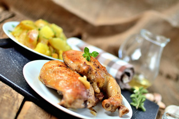 Golden grilled chicken legs on a plate and roasted potatoes, rosemary, jug with oil, oregano, salt, pepper and garlic on a wooden table