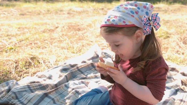 Little cute girl eats sandwich with bread and processed cheese. Family picnic. Licks her dirty fingers.