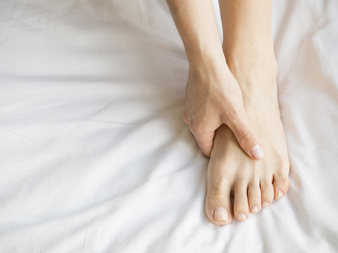 Woman Holding her Tired Feet in Hand Sitting on Bed with White Sheets.