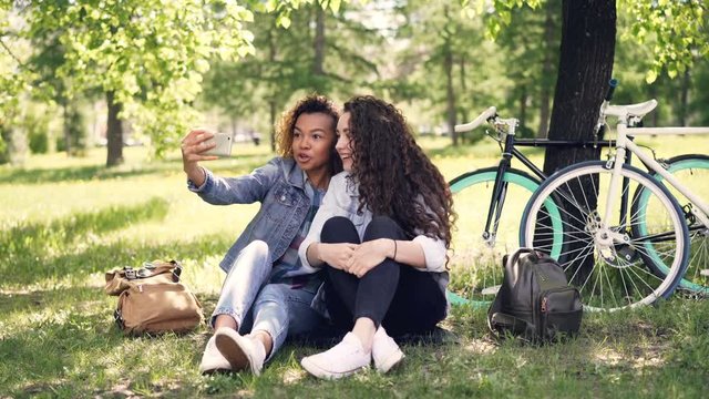 Cheerful women are looking at smartphone screen and laughing then taking selfie together posing for camera sitting on grass in park. Bicycles and backpacks are visible.