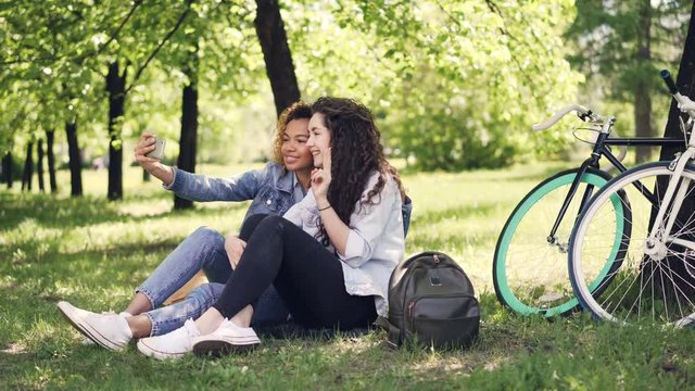 Young woman and her African American friend are taking selfie, posing and having fun sitting on lawn in park. Warm sunny day, beautiful nature and modern lifestyle concept.