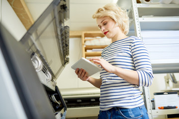 Attractive young woman in casual outfit standing near plotter in printing office and using tablet. 
