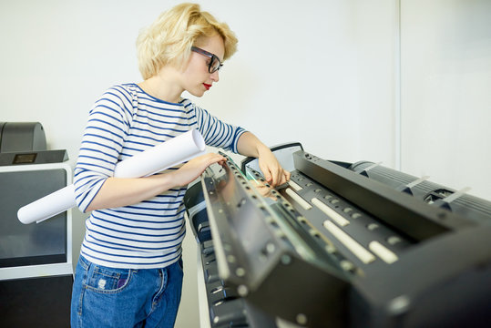Beautiful young woman in casual outfit holding roll of paper and inspecting professional plotter in printing office. 